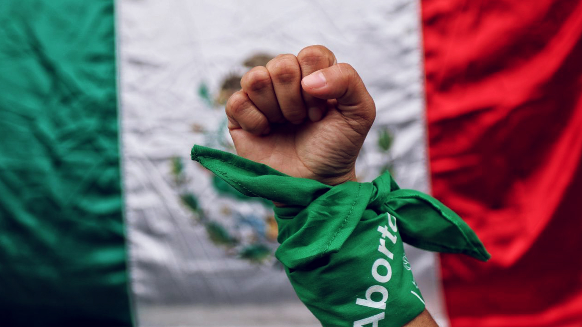 una mano con un pañuelo se levanta delante de la bandera de México en pro del aborto